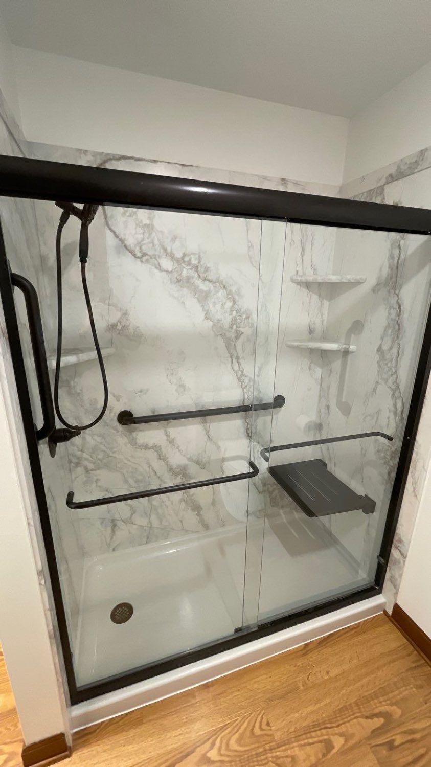 Walk-in shower with marble-look panels, black sliding glass door, grab bars, and a fold-down seat